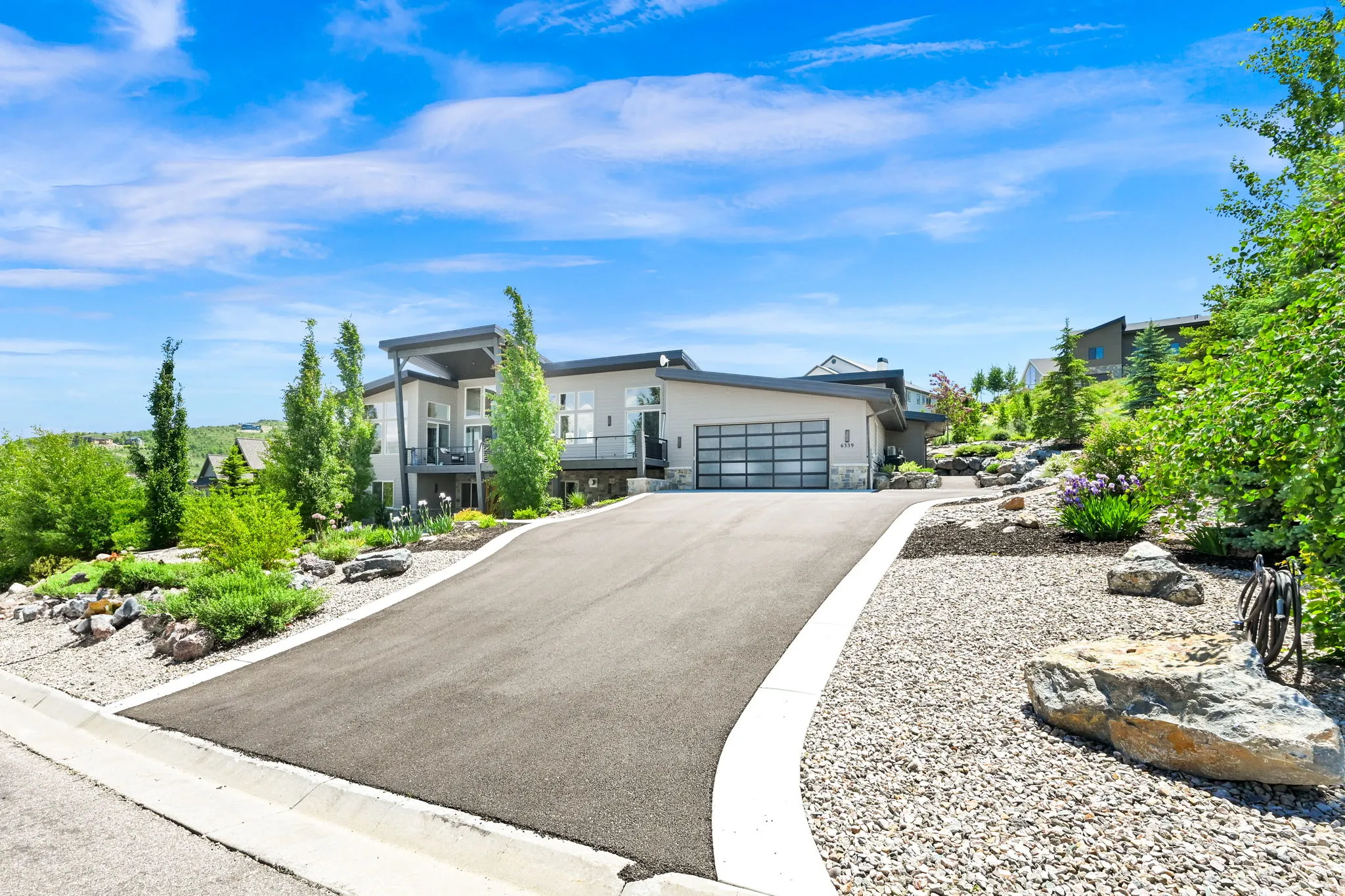 Contemporary architectural design featuring a two-car garage with a modern glass door, a paved driveway, and mature landscaping with rock features