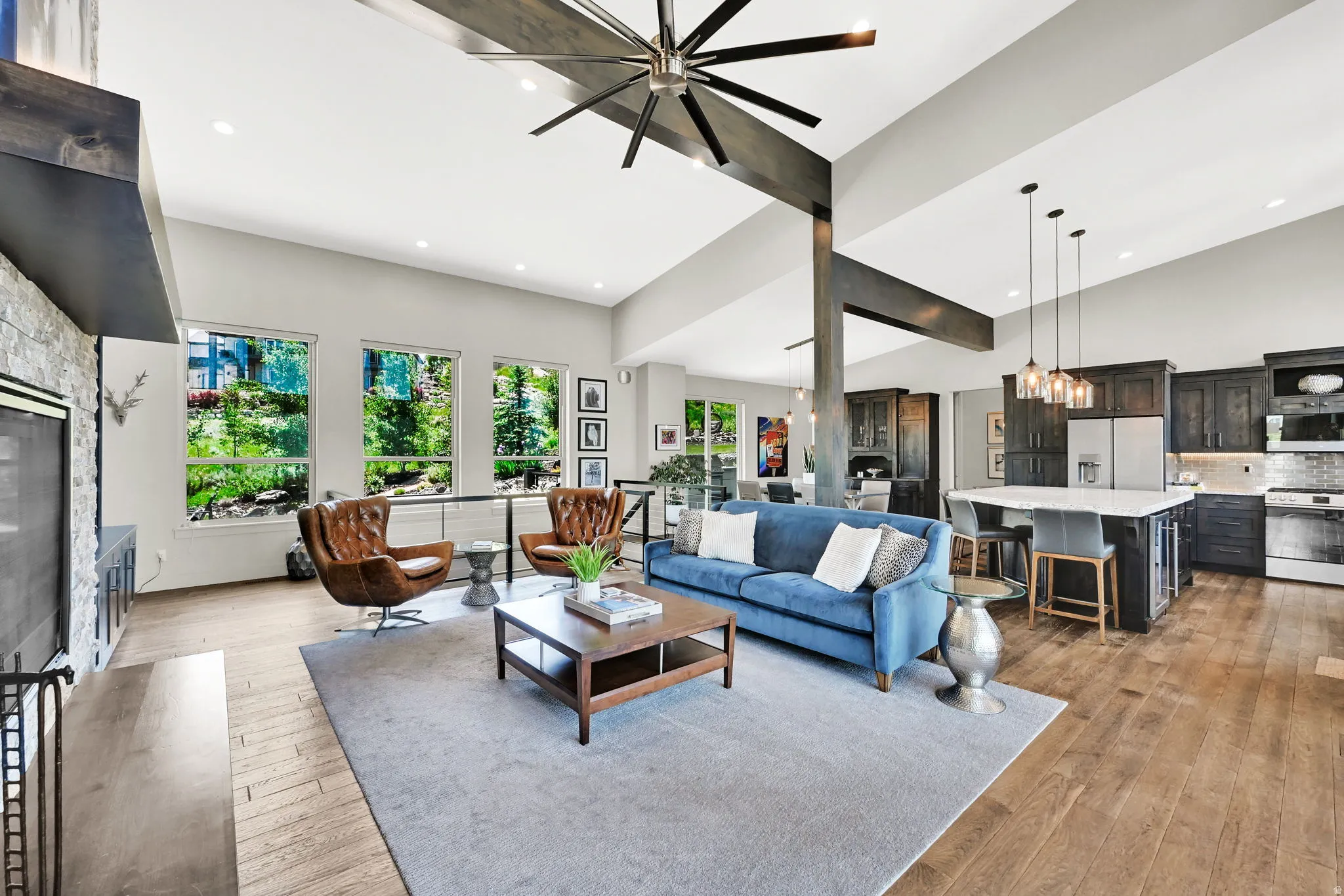 Spacious open-concept living area featuring vaulted ceilings, wood-finish flooring, a stone-front fireplace, and large windows
