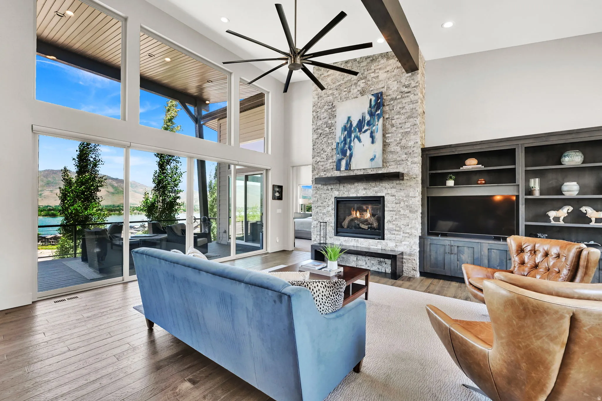 Spacious living area featuring floor-to-ceiling windows, a stone-clad fireplace, built-in shelving, wood-finish flooring, and a large ceiling fan