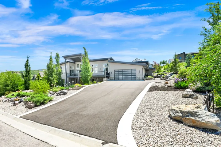 Contemporary architectural design featuring a two-car garage with a modern glass door, a paved driveway, and mature landscaping with rock features