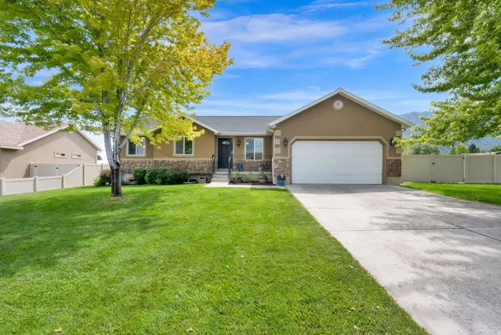 Stucco and stone facade with an attached two-car garage, concrete driveway, and a fenced yard featuring mature trees and a manicured lawn