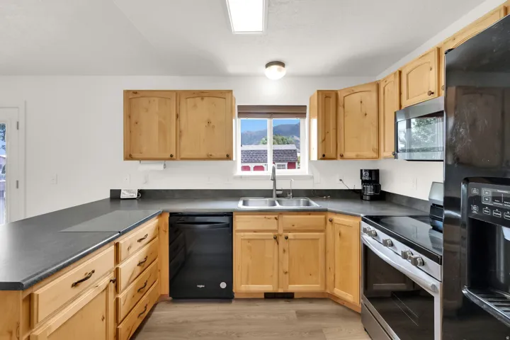 Kitchen featuring natural wood cabinetry, dark countertops, a double basin stainless steel sink, black and stainless steel appliances, and wood-finish flooring