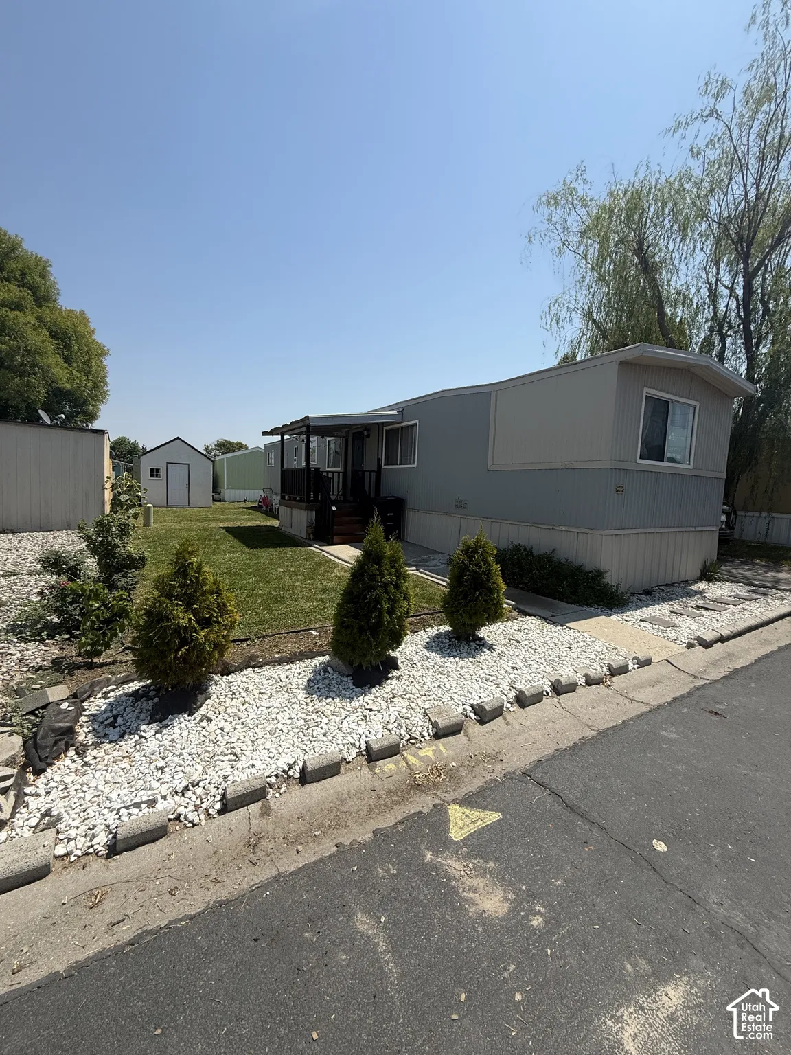 Charming manufactured home featuring a manicured front landscape with mature evergreen plantings, white gravel hardscaping, and defined border edging