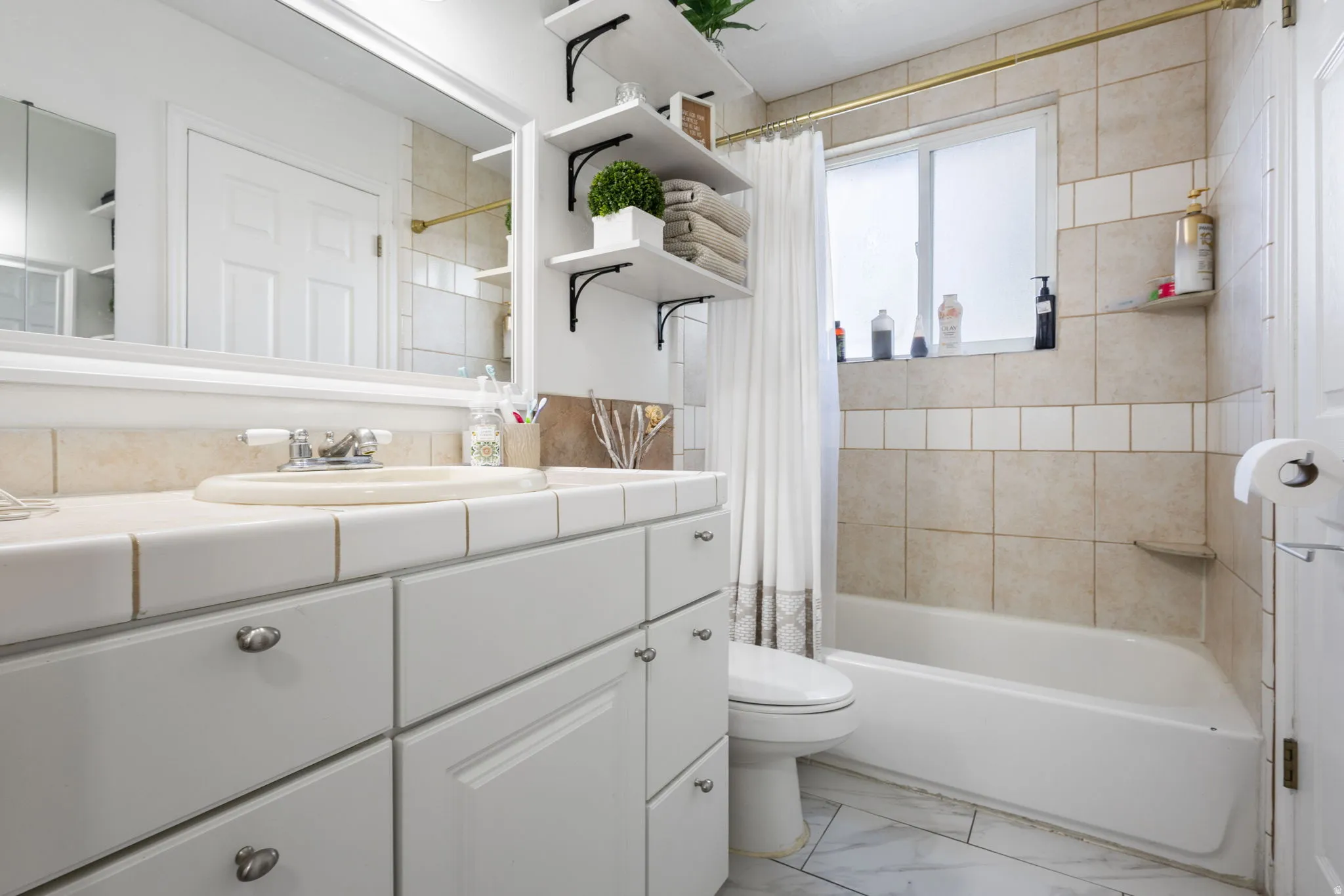 Vanity with integrated sink and tile countertop, white cabinetry with silver hardware, large framed mirror, built-in shelving, tub with tiled surround, and a frosted window