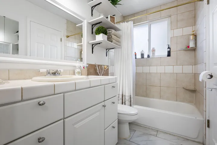 Vanity with integrated sink and tile countertop, white cabinetry with silver hardware, large framed mirror, built-in shelving, tub with tiled surround, and a frosted window