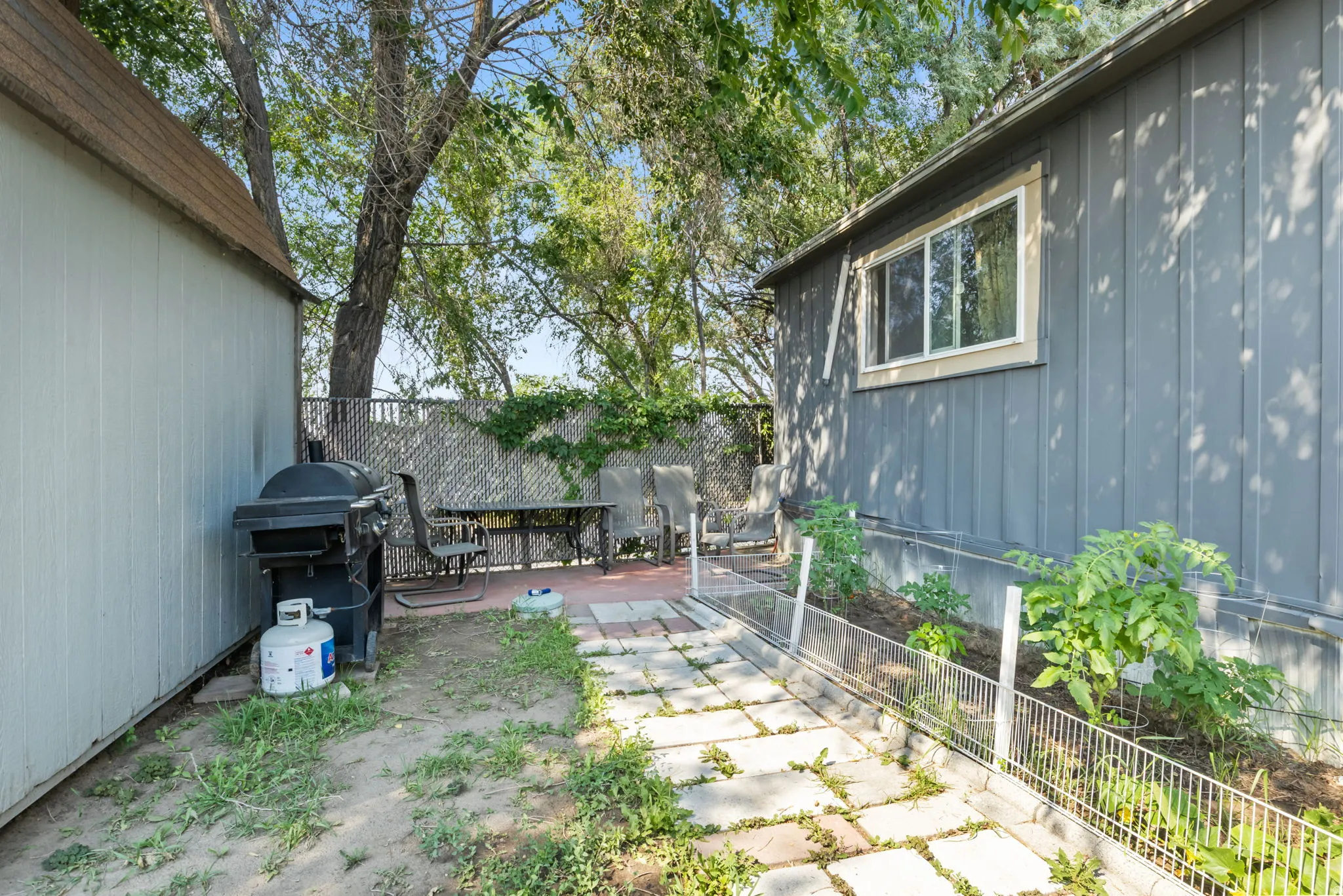 Outdoor space featuring a grey-sided structure with a white-trimmed window, a separate utility shed, a paver patio area, and a garden bed