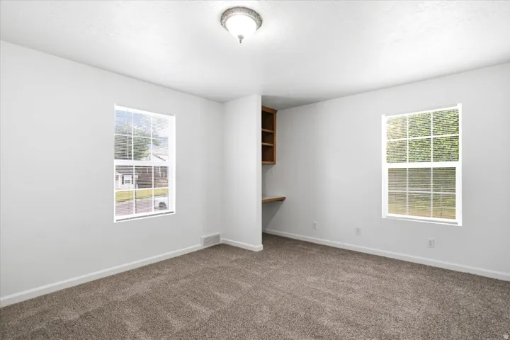 Bright interior room with plush carpeting, two windows, and a built-in wooden desk with shelving