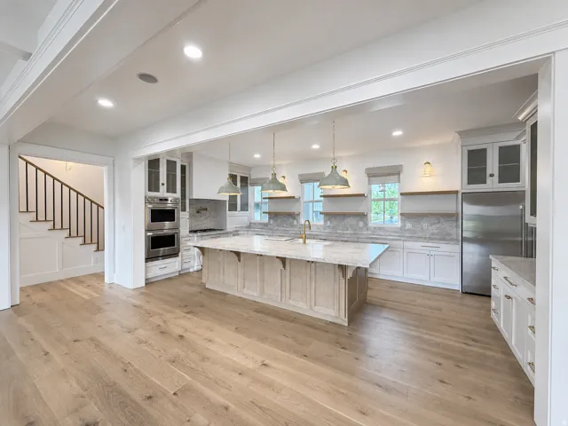 Open-concept kitchen featuring a large central light wood base island with Taj Mahal leather quartzite stone counters, stainless steel appliances, white cabinetry, and light engineered wood flooring throughout.
