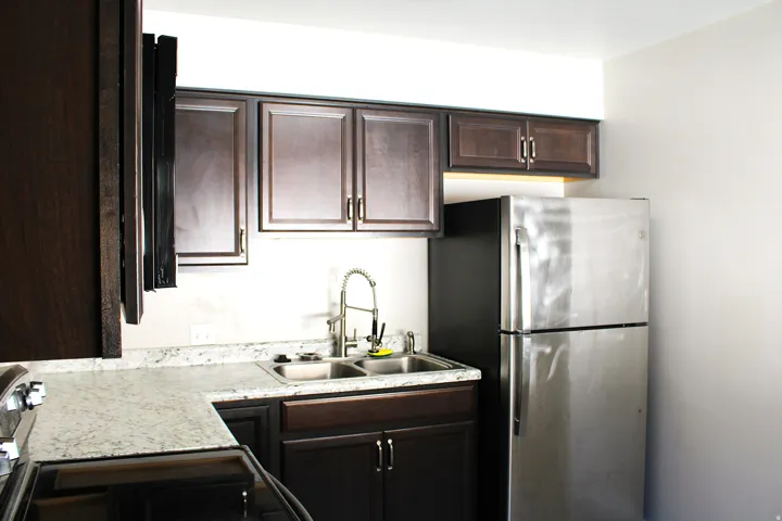 Kitchen featuring dark wood-finish cabinetry, light-toned countertops, a double basin stainless steel sink with a gooseneck faucet, and a stainless steel refrigerator