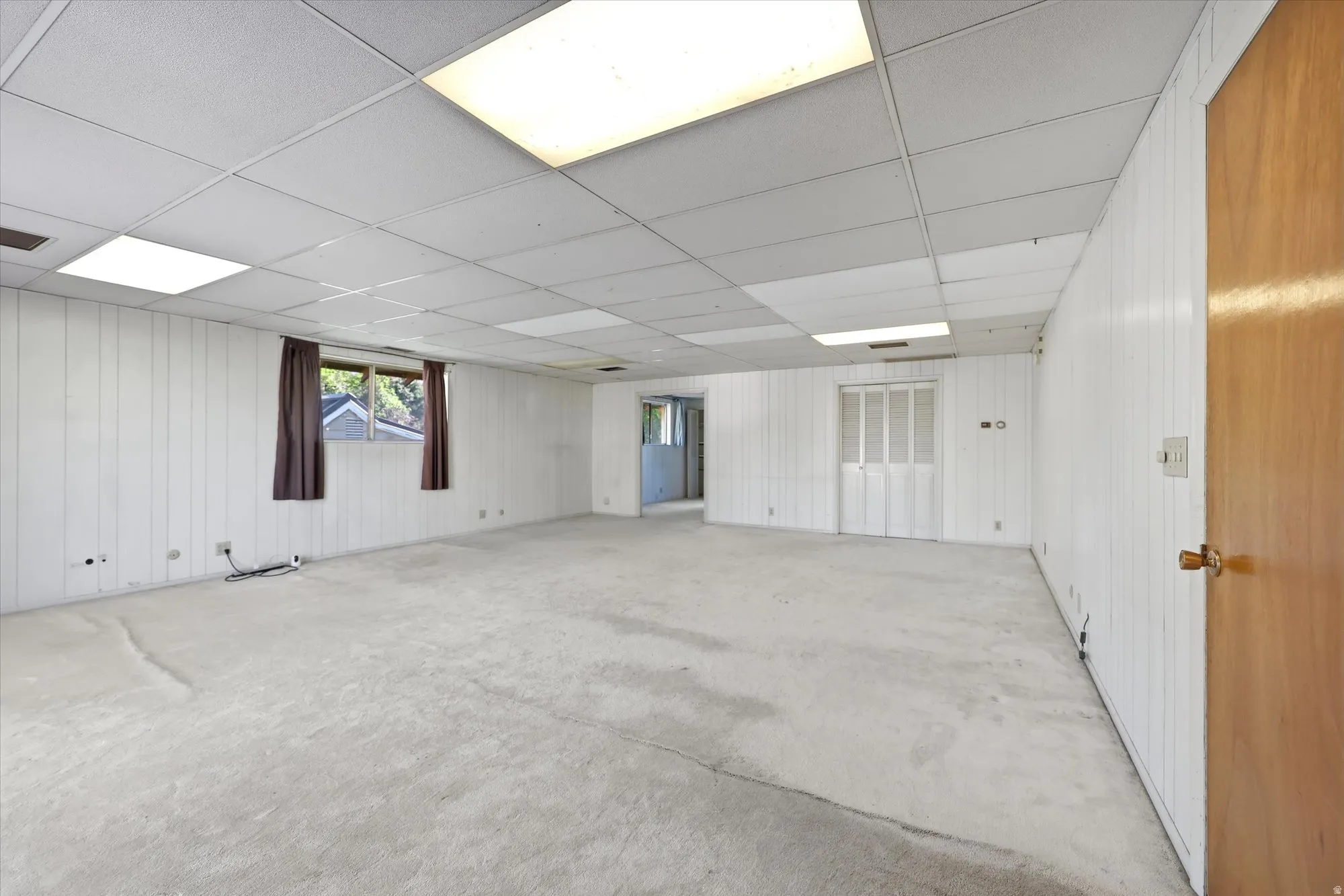 Spacious room featuring light-colored carpet, white vertical paneling, and a drop ceiling with integrated lighting