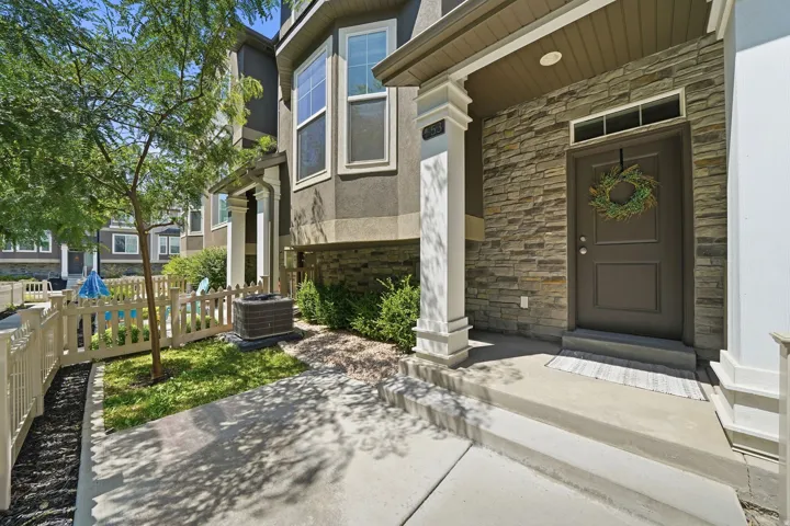 Front exterior featuring a stone-clad entry, dark gray door with transom window, concrete porch with decorative pillars, and a picket-fenced yard with mature landscaping