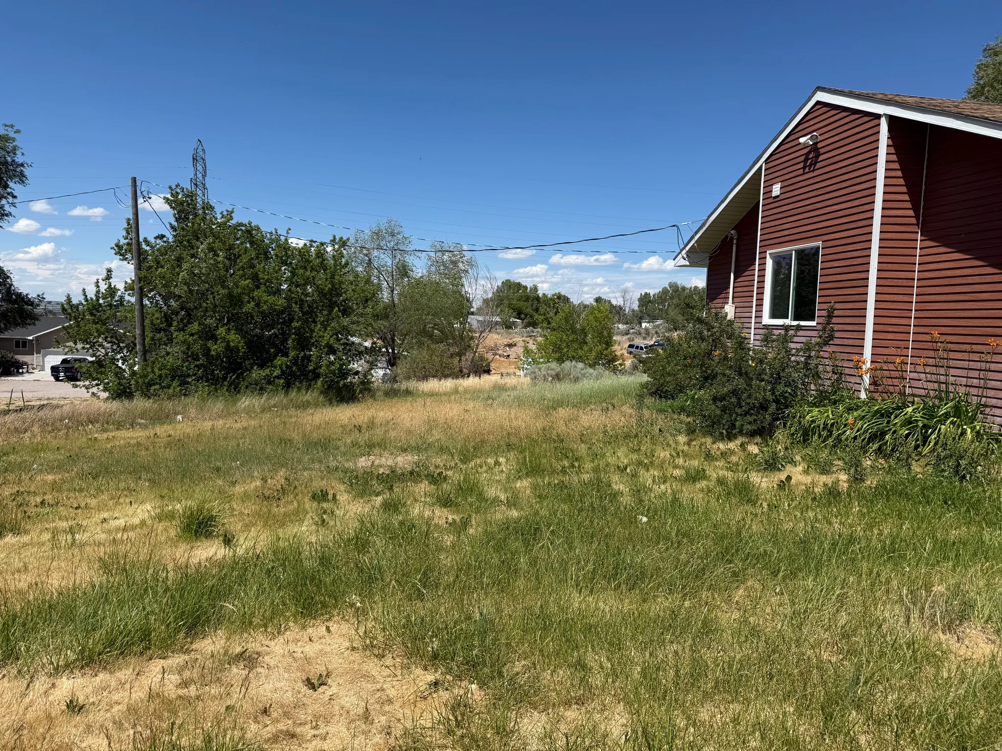 Expansive grassy lot featuring a red siding structure with white trim, a visible window, and mature plantings