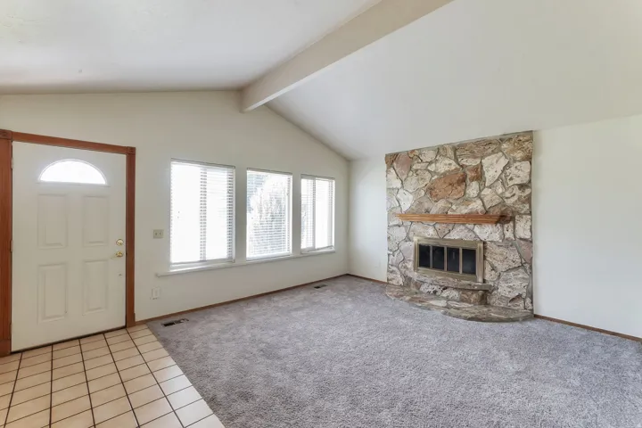 Vaulted ceilings enhance this living space, featuring a stone fireplace with a wood mantle, multiple windows, and a tiled entry