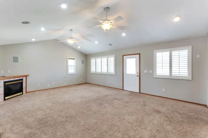 Spacious living area featuring a vaulted ceiling with recessed lighting and ceiling fans, a fireplace with a tile surround and wood mantel, multiple windows with white plantation shutters, and a rear exit door with a clear glass insert