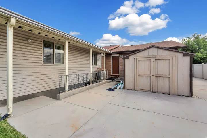 Paved concrete patio area featuring covered porch with support columns, metal railing, and an exterior storage shed
