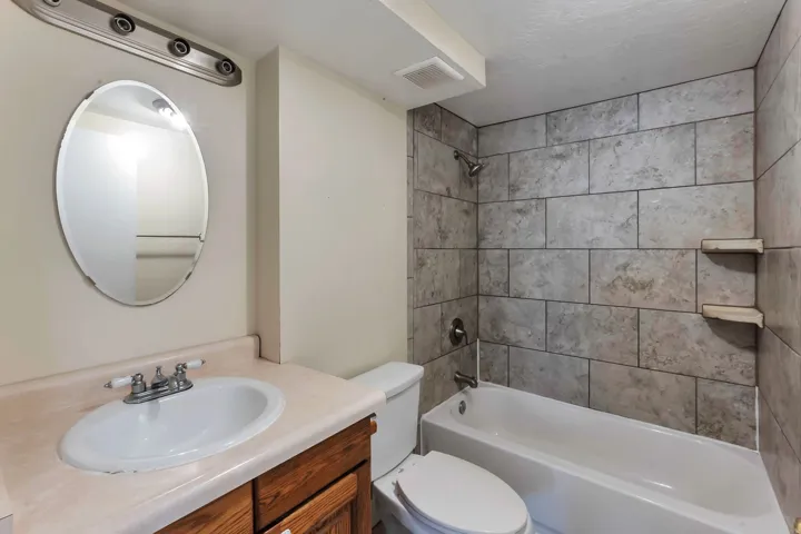 Bathroom featuring a white bathtub, ceramic tile shower surround, wood-finish vanity with an integrated sink, oval mirror, and overhead lighting