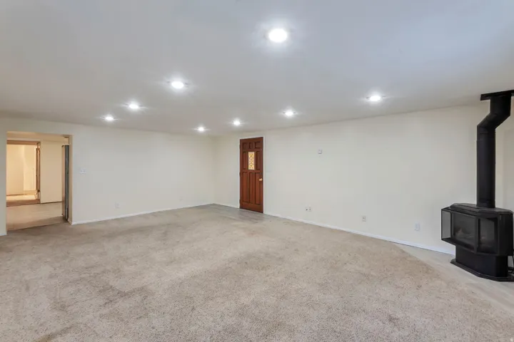 Spacious room featuring neutral carpeting, recessed lighting, a freestanding wood-burning stove with a black flue, a wooden entry door with decorative glass, and a wide interior doorway