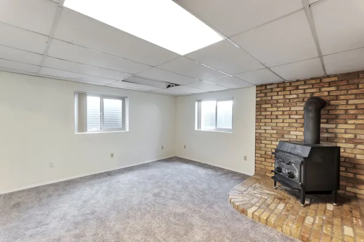 Spacious room featuring a wood-burning stove on a brick hearth, a full brick accent wall, two windows with blinds, light-toned carpeting, and a drop ceiling with recessed lighting