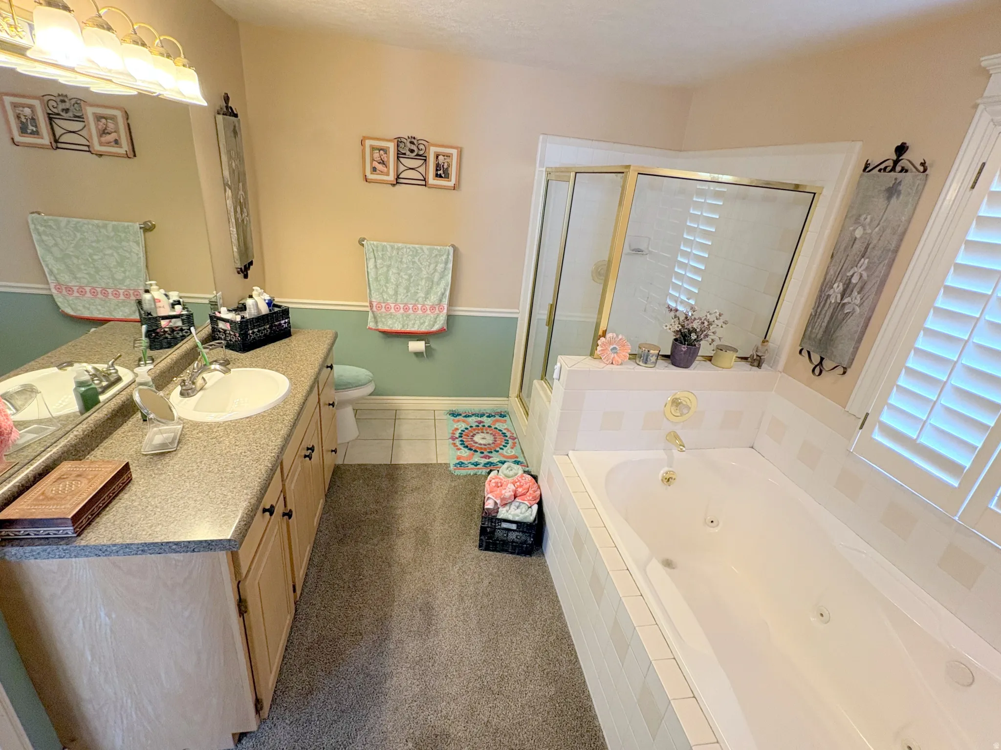 Bathroom featuring a built-in jetted tub, a separate glass-enclosed shower, a double vanity with a speckled countertop, light wood cabinetry, and plantation shutters