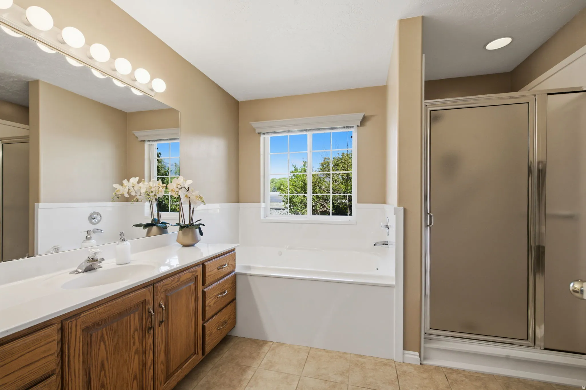 Bathroom featuring a wood-finish double vanity with white countertops, a built-in tub with a window, a separate glass-enclosed shower, and tiled flooring