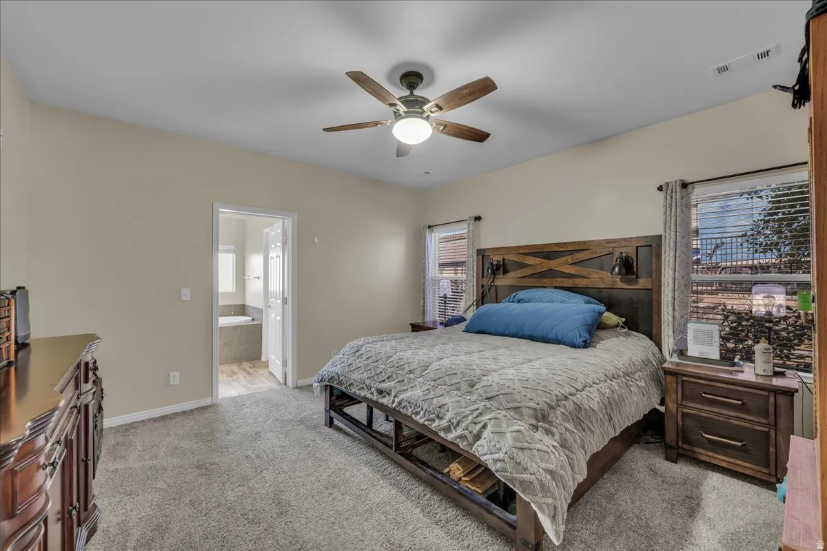 Carpeted bedroom featuring a vaulted ceiling with a ceiling fan, two windows with blinds, and a doorway opening to a bathroom with a built-in tub