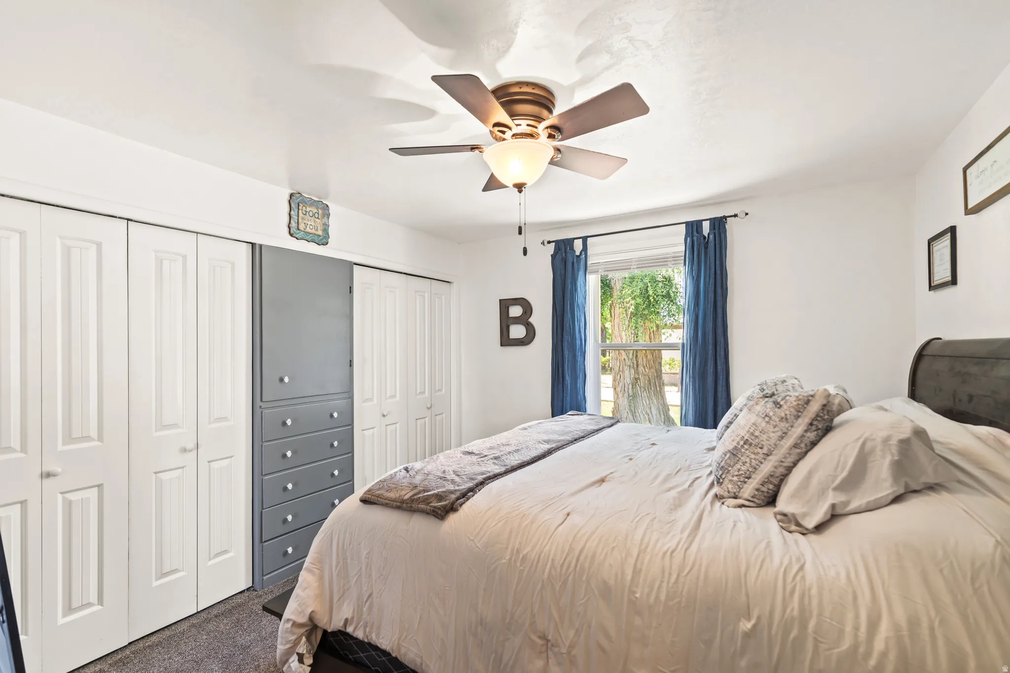 Bedroom featuring a window with exterior tree visibility, ceiling fan light fixture, two sets of white bi-fold closet doors, and a built-in gray drawer and cabinet unit