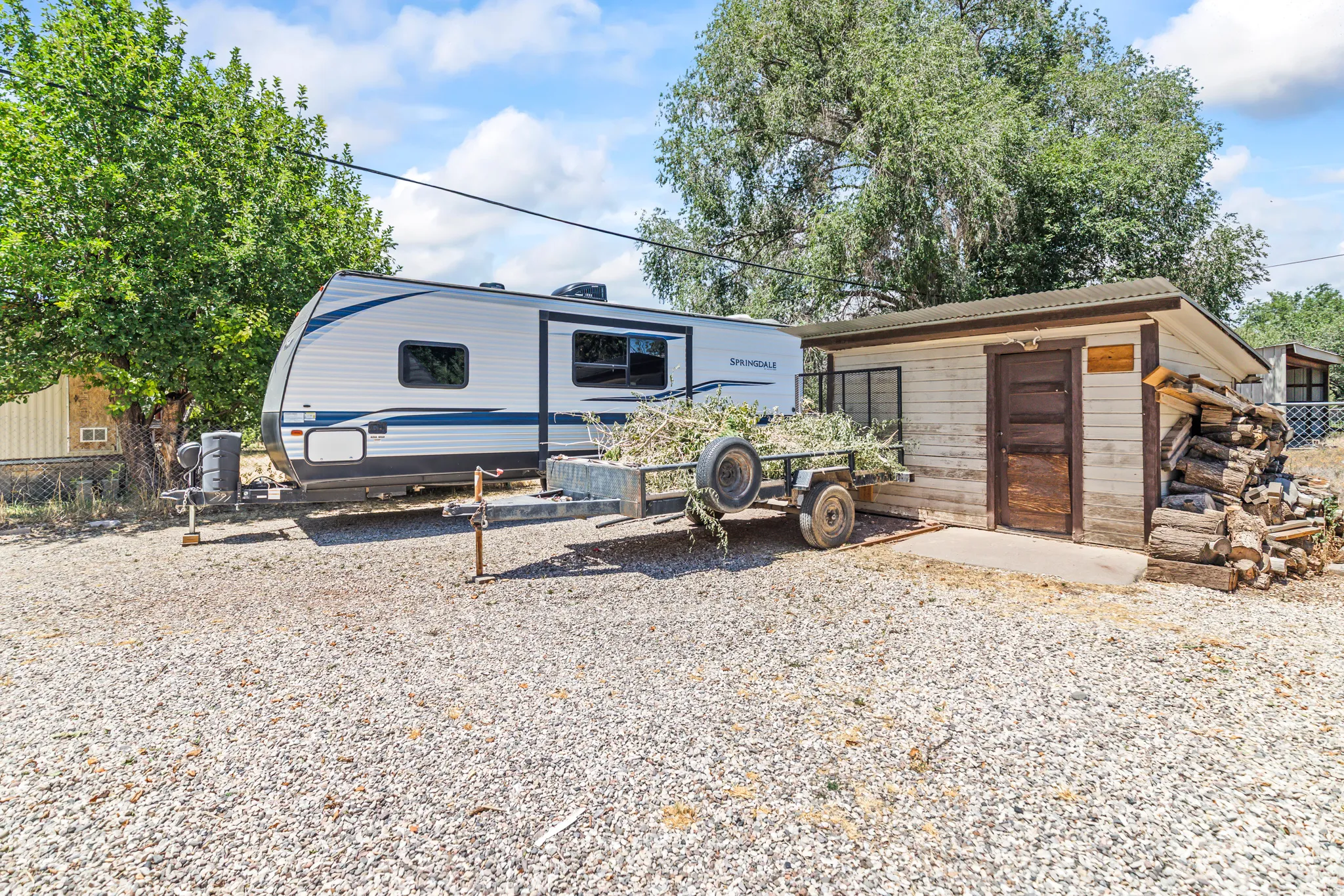 Gravel lot featuring a detached shed with wood siding and a corrugated metal roof, complemented by mature trees in the background