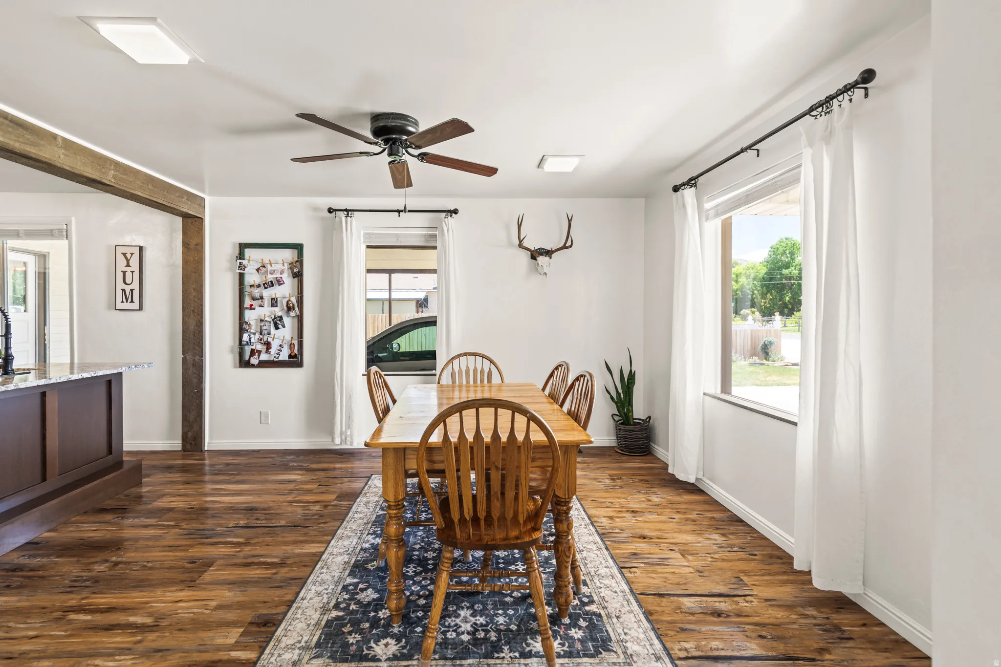 Spacious dining area featuring wood-finish flooring, two large windows with black curtain rods, a ceiling fan with integrated lighting, and exposed wooden beams defining the open concept