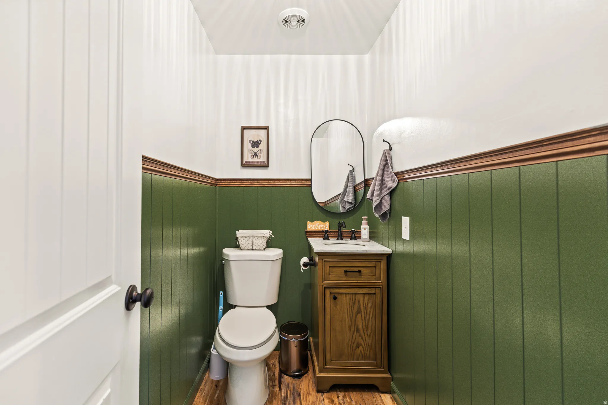Powder room featuring vertical paneling, a wood-finish vanity with an integrated sink, a dark bronze faucet, and an oval wall mirror