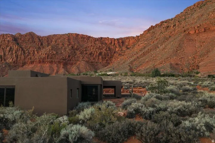 Modern-style exterior with a flat roofline, stucco finish, and dark-framed windows, surrounded by natural desert landscaping and dramatic red rock formations