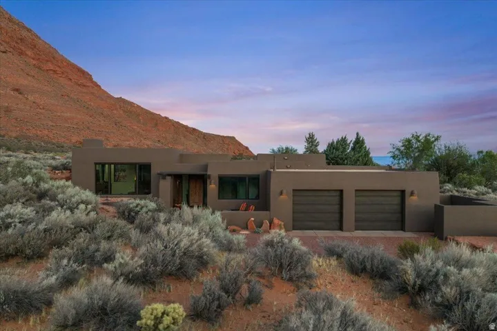 Southwestern-style residence featuring a stucco exterior, integrated two-car garage with dark wood-finish doors, prominent rock formations, and arid landscaping