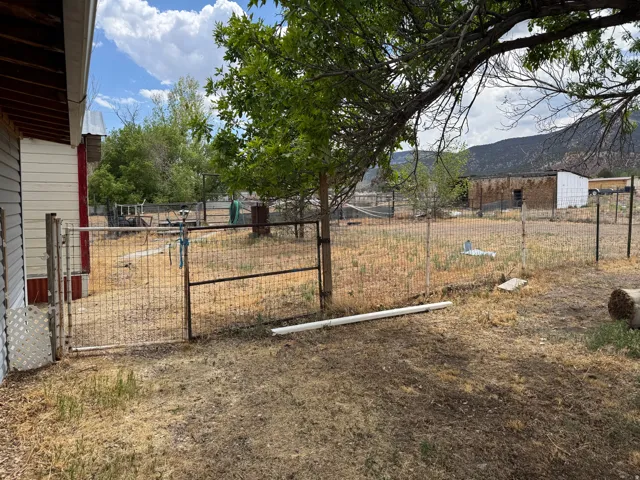 Fenced yard area featuring a metal gate and mature trees, with a distant building and mountainous backdrop