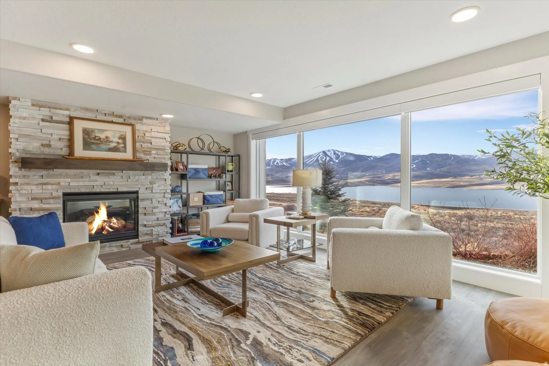 Living area featuring a stone-clad fireplace with a wood mantel, expansive windows showcasing mountain and lake scenery, recessed lighting, and wood-finish flooring