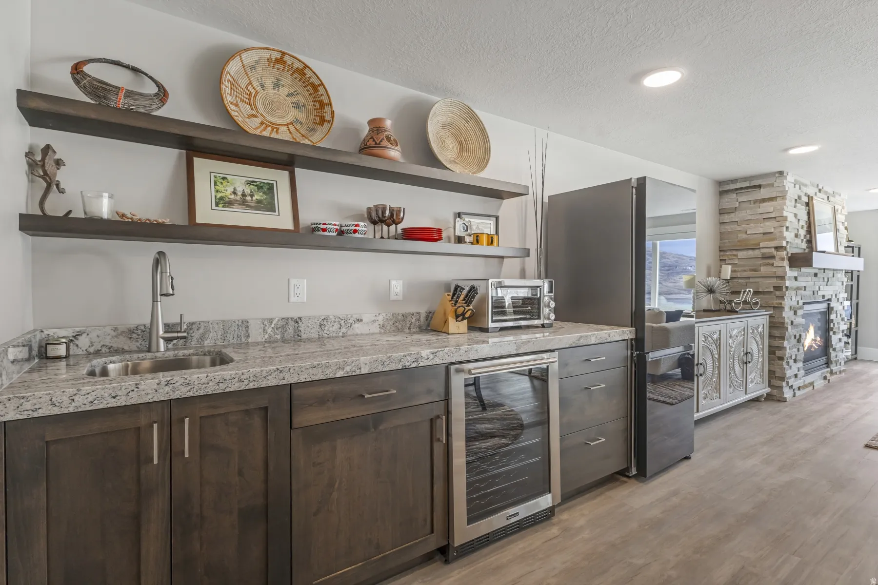 Wet bar featuring stone-finish countertops, undermount sink with a gooseneck faucet, dark wood cabinetry, and open shelving