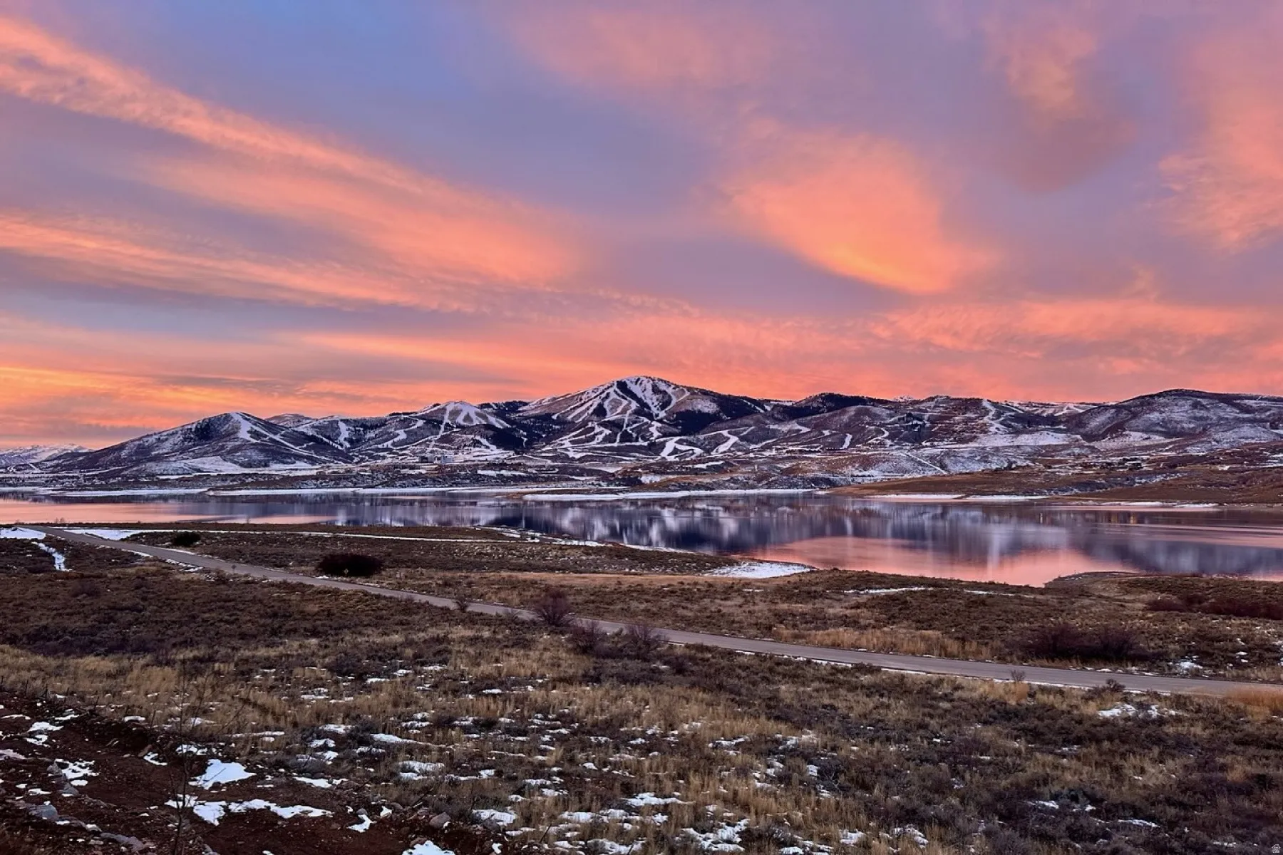 Expansive mountain landscape featuring a large body of water reflecting the colorful sky, with snow-dusted peaks in the background