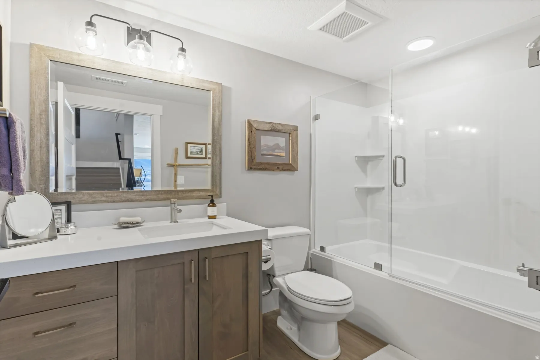Modern bathroom featuring a wood-finish vanity with a white countertop and undermount sink, large framed mirror, glass-enclosed bathtub with a shower, and wood-finish flooring
