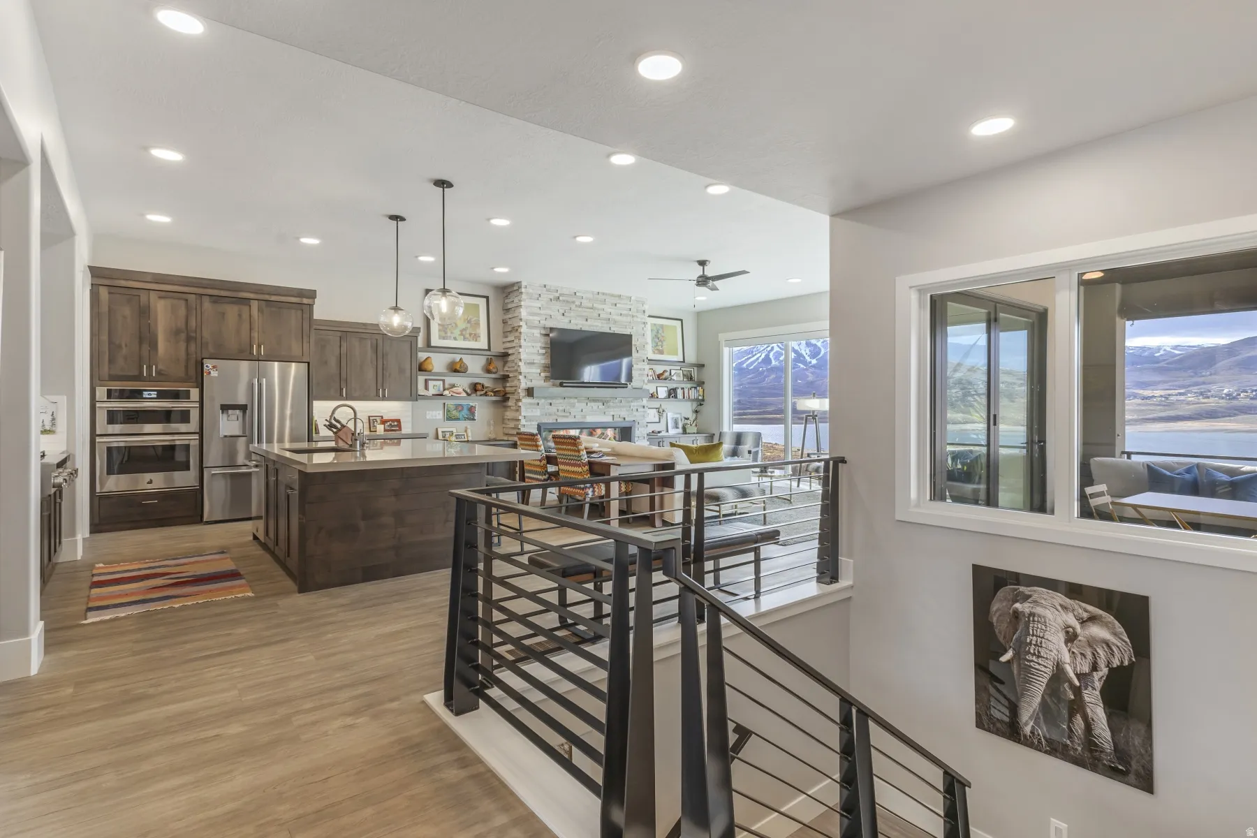 Open-concept living space with wood-finish flooring, a kitchen island, stainless steel appliances, a stone accent wall, and a large window overlooking a body of water and mountains