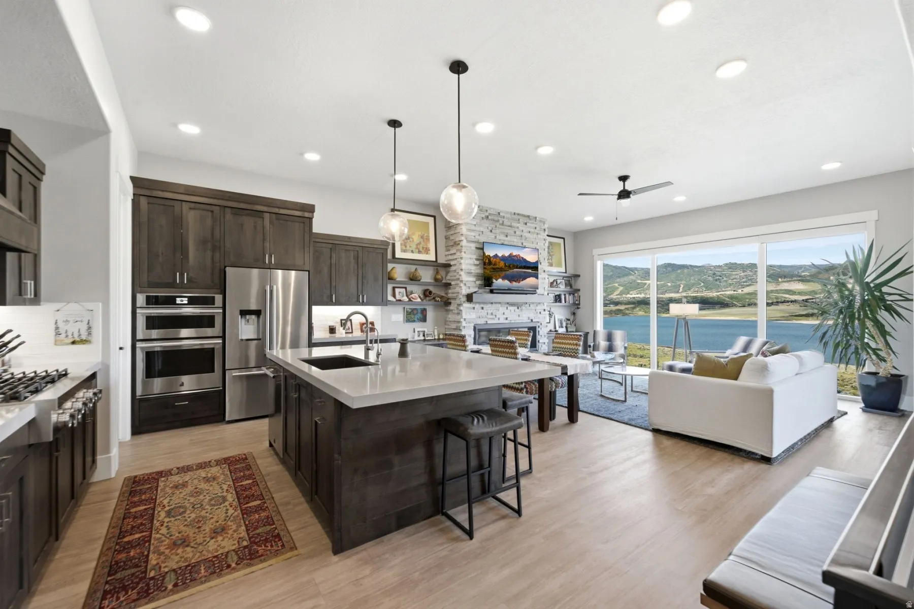 Open-concept living area featuring a kitchen with dark wood cabinetry and stainless steel appliances, a center island with pendant lighting, a living room with a stone fireplace, and expansive sliding glass doors overlooking a body of water and distant hi