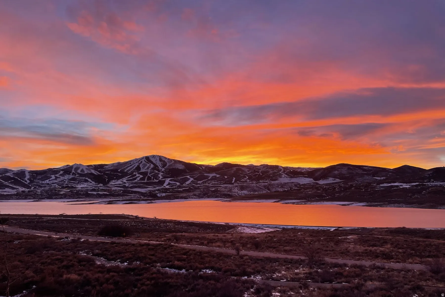 Expansive landscape with a lake reflecting an orange and pink sky, framed by snow-dusted mountains