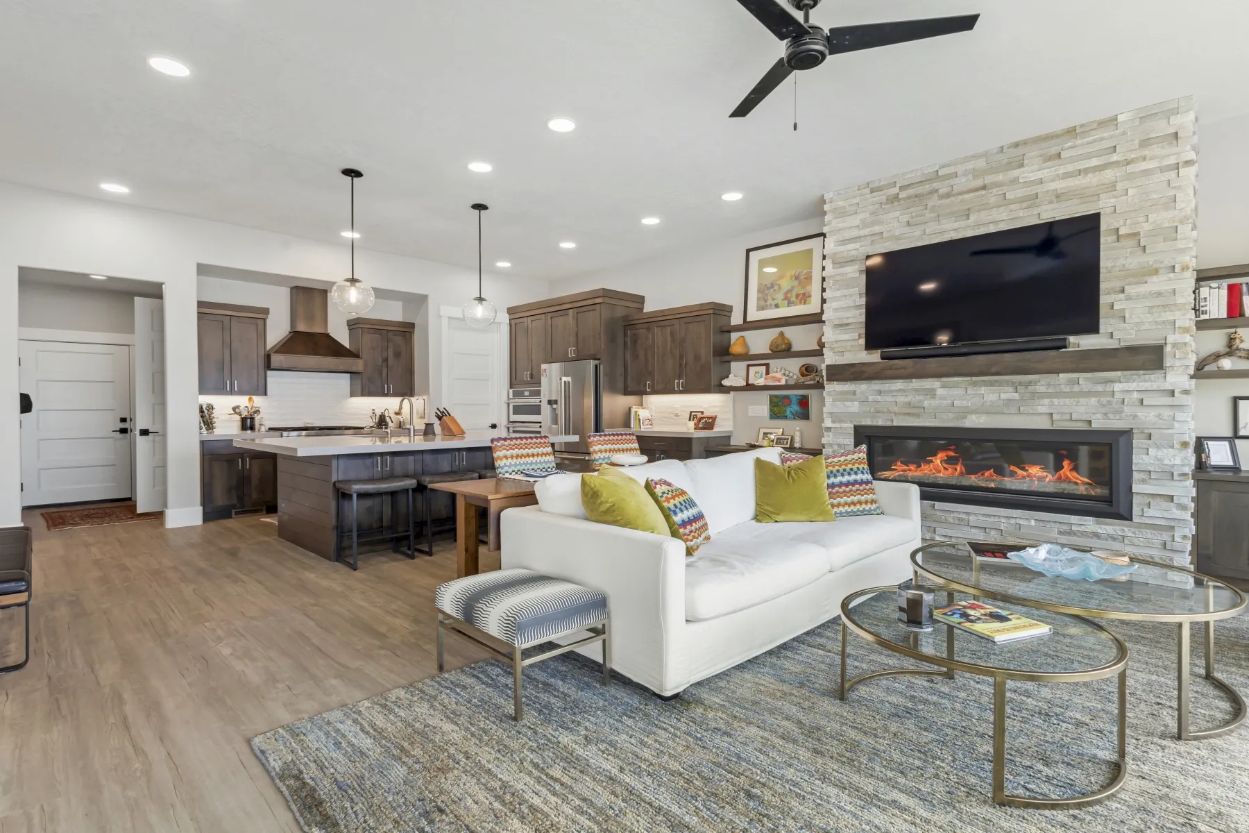 Open-concept layout featuring wood-finish flooring, a stacked stone fireplace with a wood mantle, and a kitchen island with pendant lighting