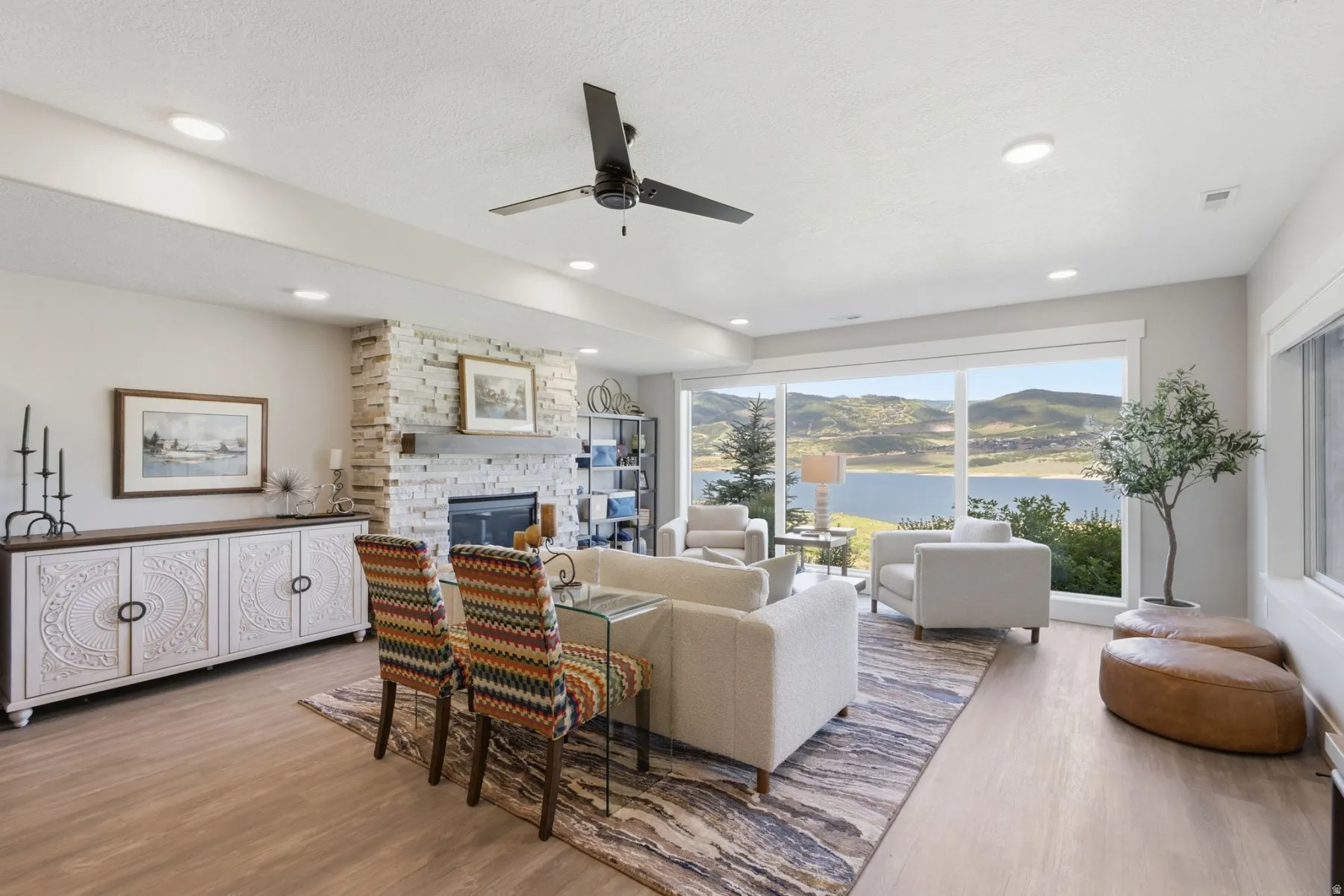 Spacious living area featuring large windows, a stone fireplace with a wood mantle, wood-finish flooring, recessed lighting, and a ceiling fan