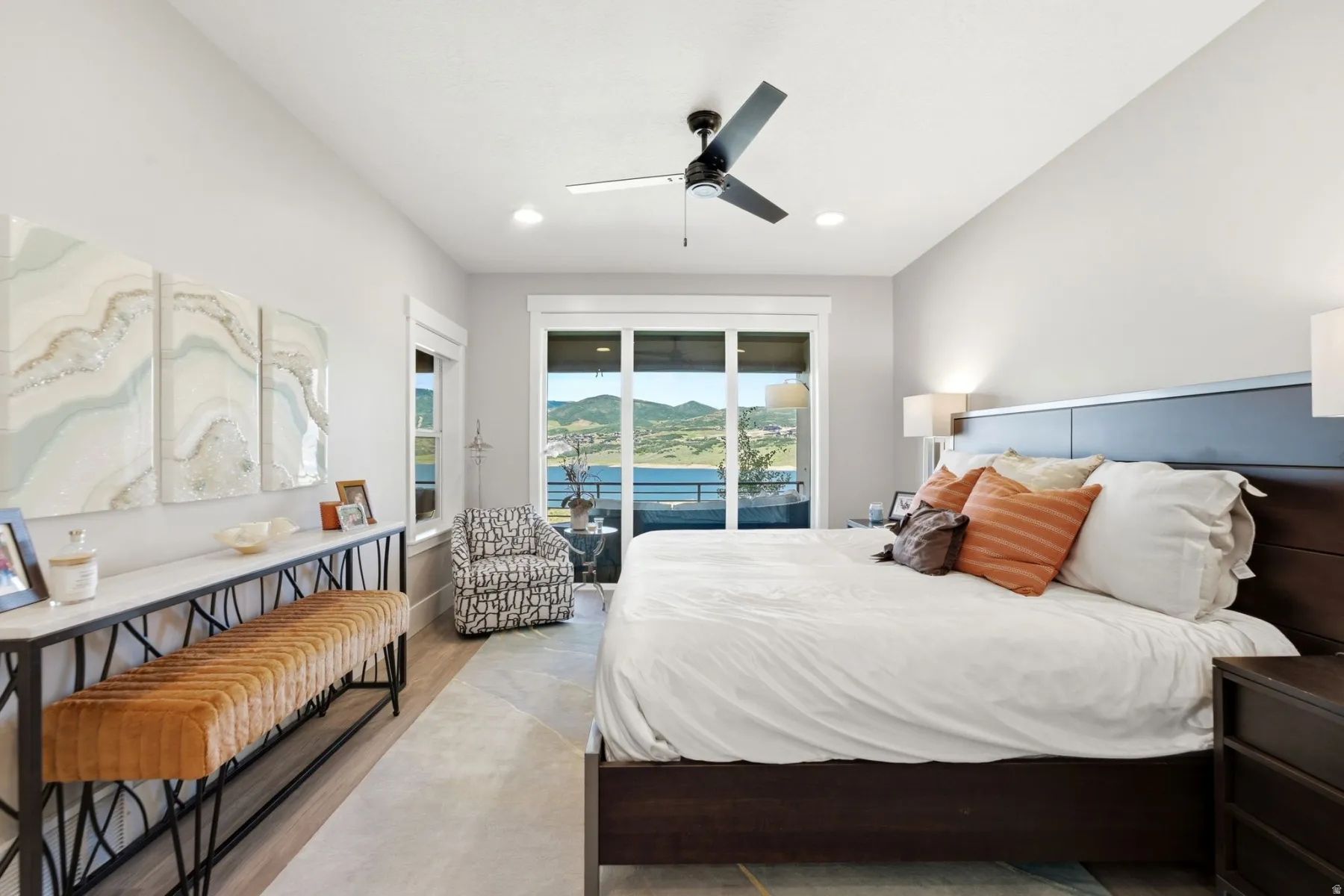 Spacious bedroom featuring wood-finish flooring, a contemporary ceiling fan with integrated lighting, and a sliding glass door opening to an outdoor area with a mountain and water backdrop