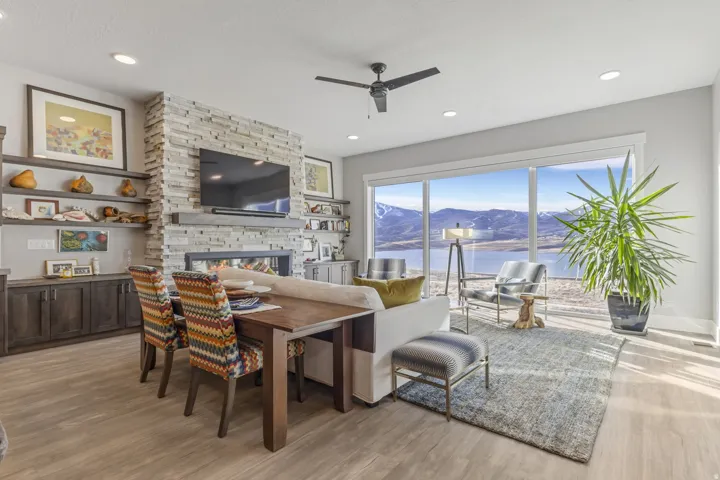 Open-concept living area featuring a stone-clad fireplace with built-in shelving, wood-finish flooring, a ceiling fan, recessed lighting, and expansive windows showcasing mountain and water scenery
