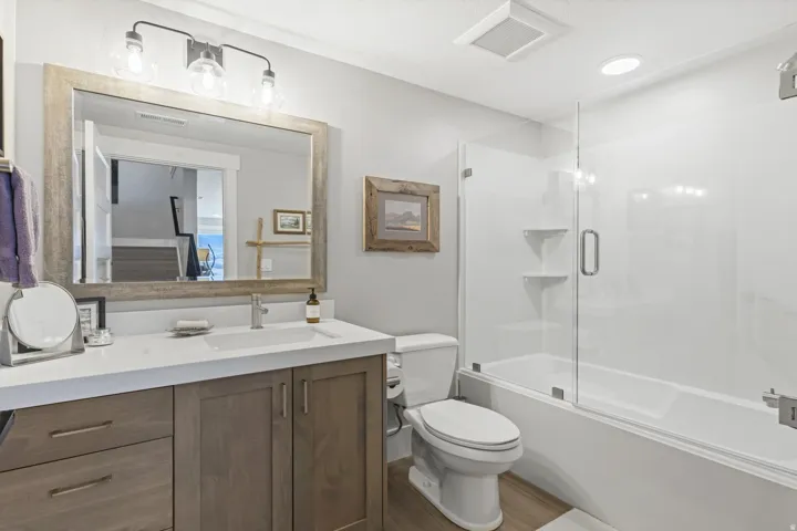 Modern bathroom featuring a wood-finish vanity with a white countertop and undermount sink, large framed mirror, glass-enclosed bathtub with a shower, and wood-finish flooring