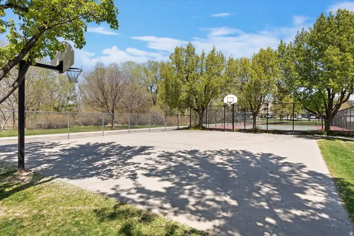 Outdoor basketball court featuring two hoops, surrounded by chain-link fencing, and mature trees providing natural shade