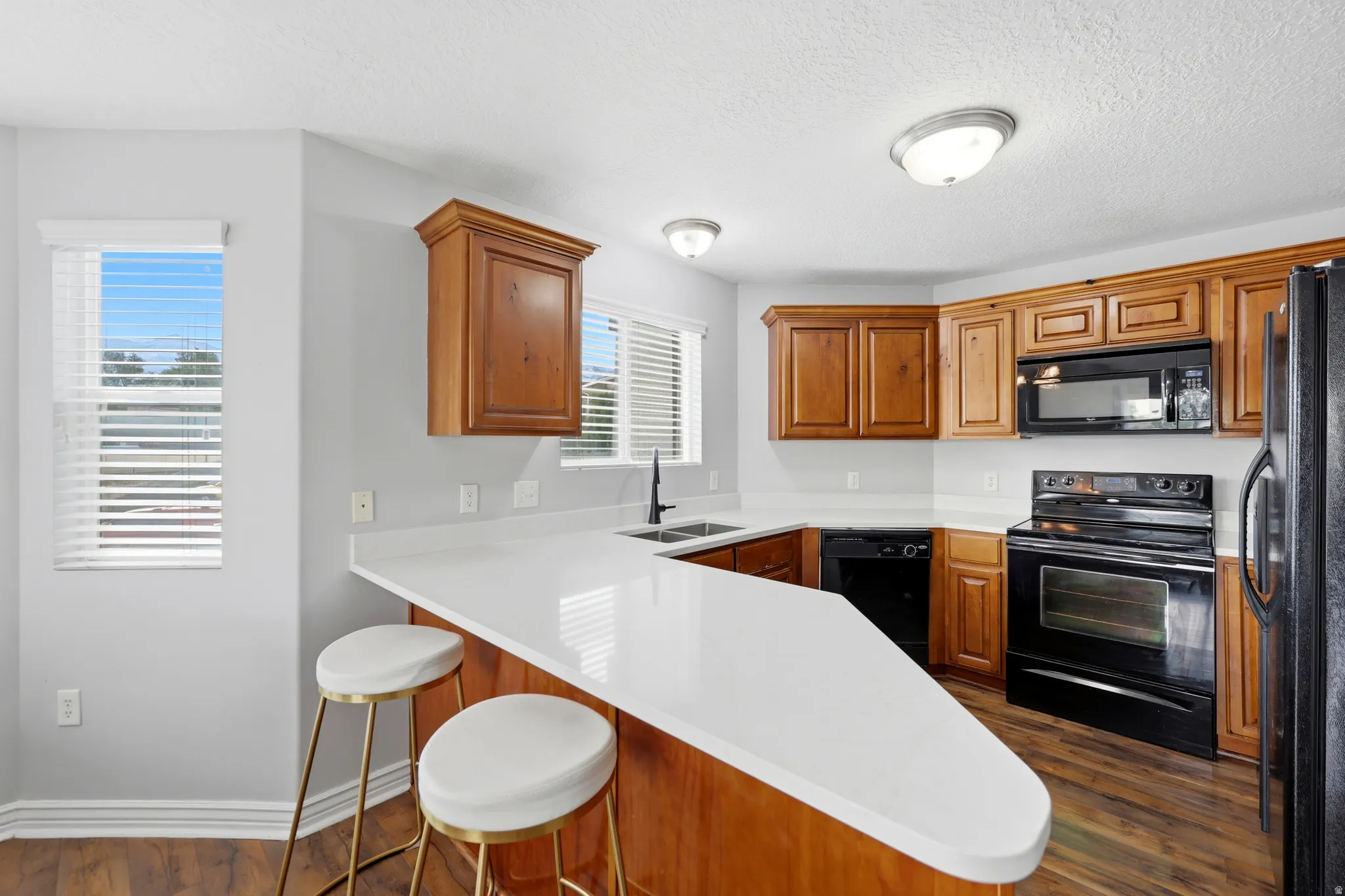 Kitchen featuring a peninsula with a white countertop, wood-finish cabinetry, black appliances, a double basin sink with a matte black faucet, and hard surface flooring