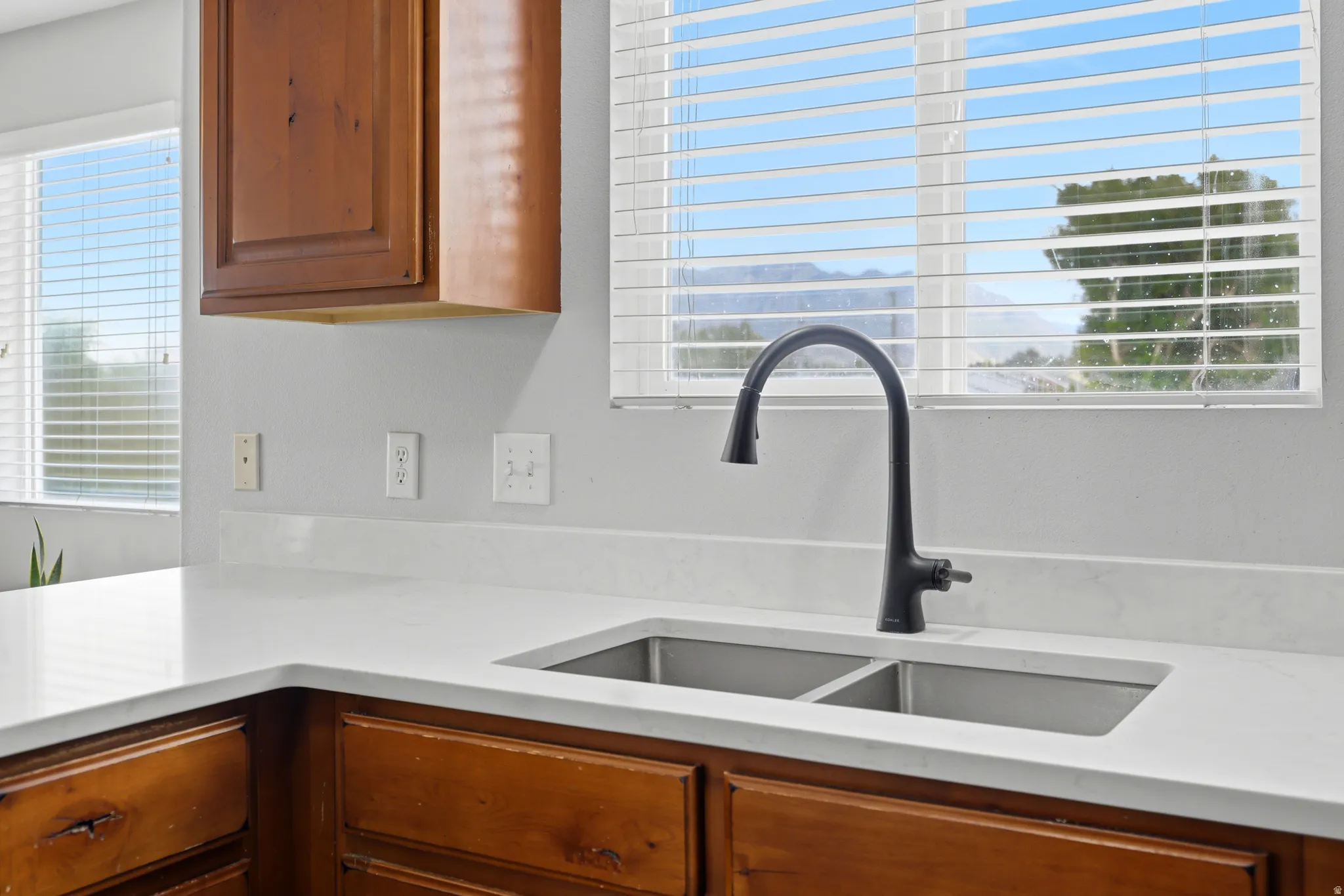 Kitchen featuring a stainless steel double basin sink with a matte black gooseneck faucet, white solid surface countertops, light wood-finish cabinetry, and a window with horizontal blinds