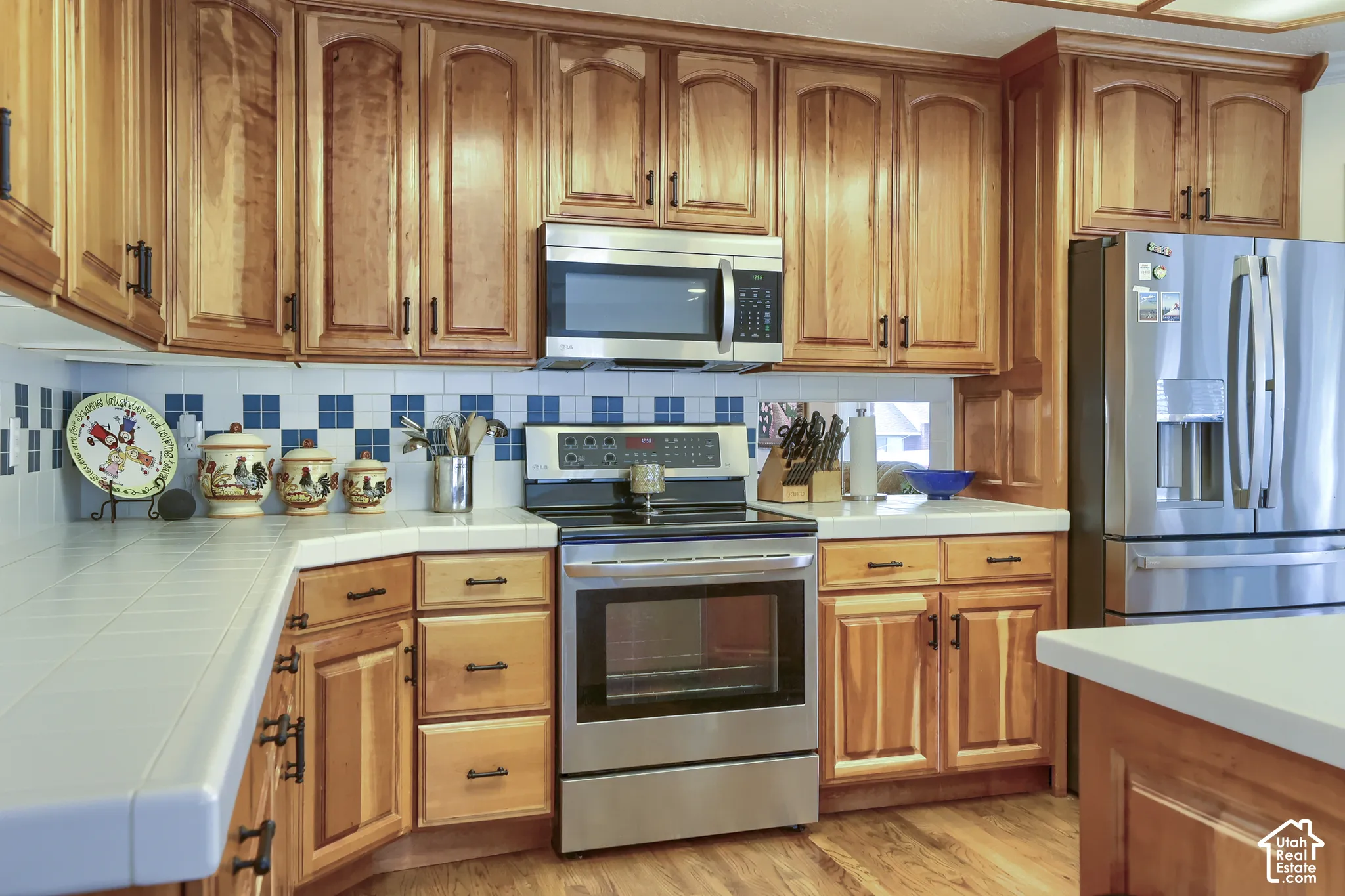 Kitchen featuring wood cabinetry, stainless steel appliances, tile countertops, a tiled backsplash, and wood-finish flooring