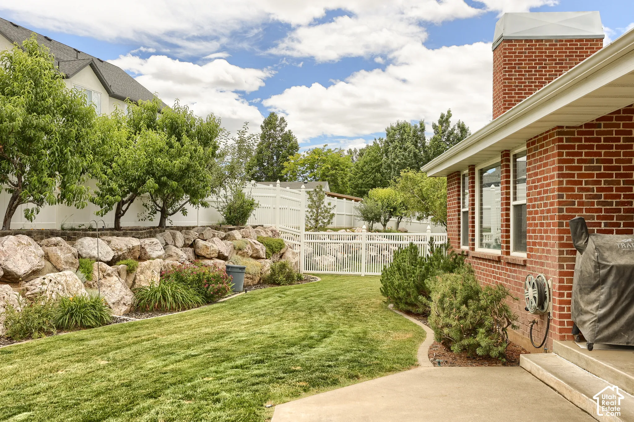 Expansive backyard featuring a lush lawn, a concrete patio, a red brick exterior, and a rock retaining wall with mature trees and shrubbery