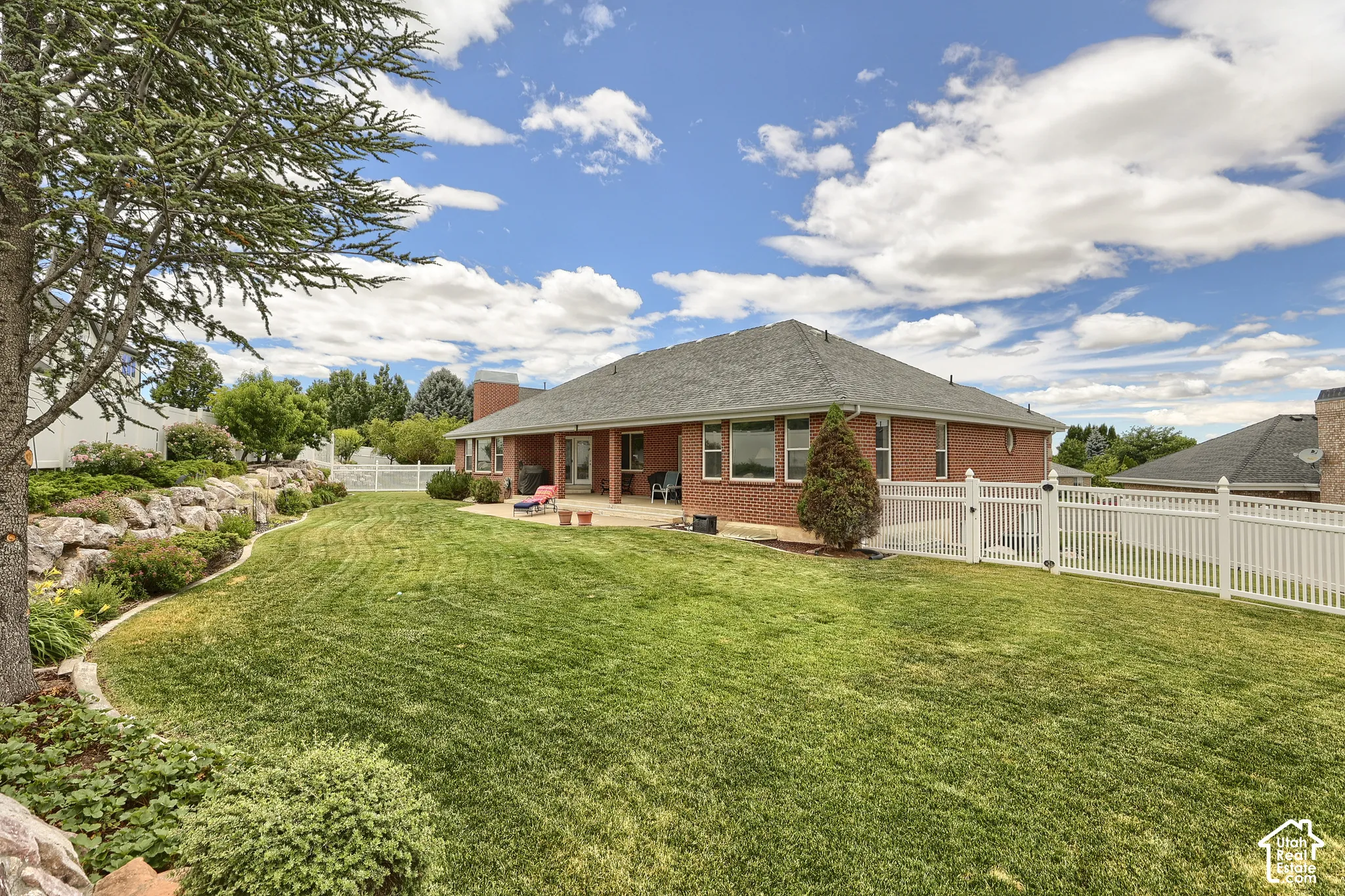 Expansive backyard featuring a manicured lawn, a white picket fence, a rock retaining wall with mature landscaping, a paved patio, and a brick exterior with multiple windows